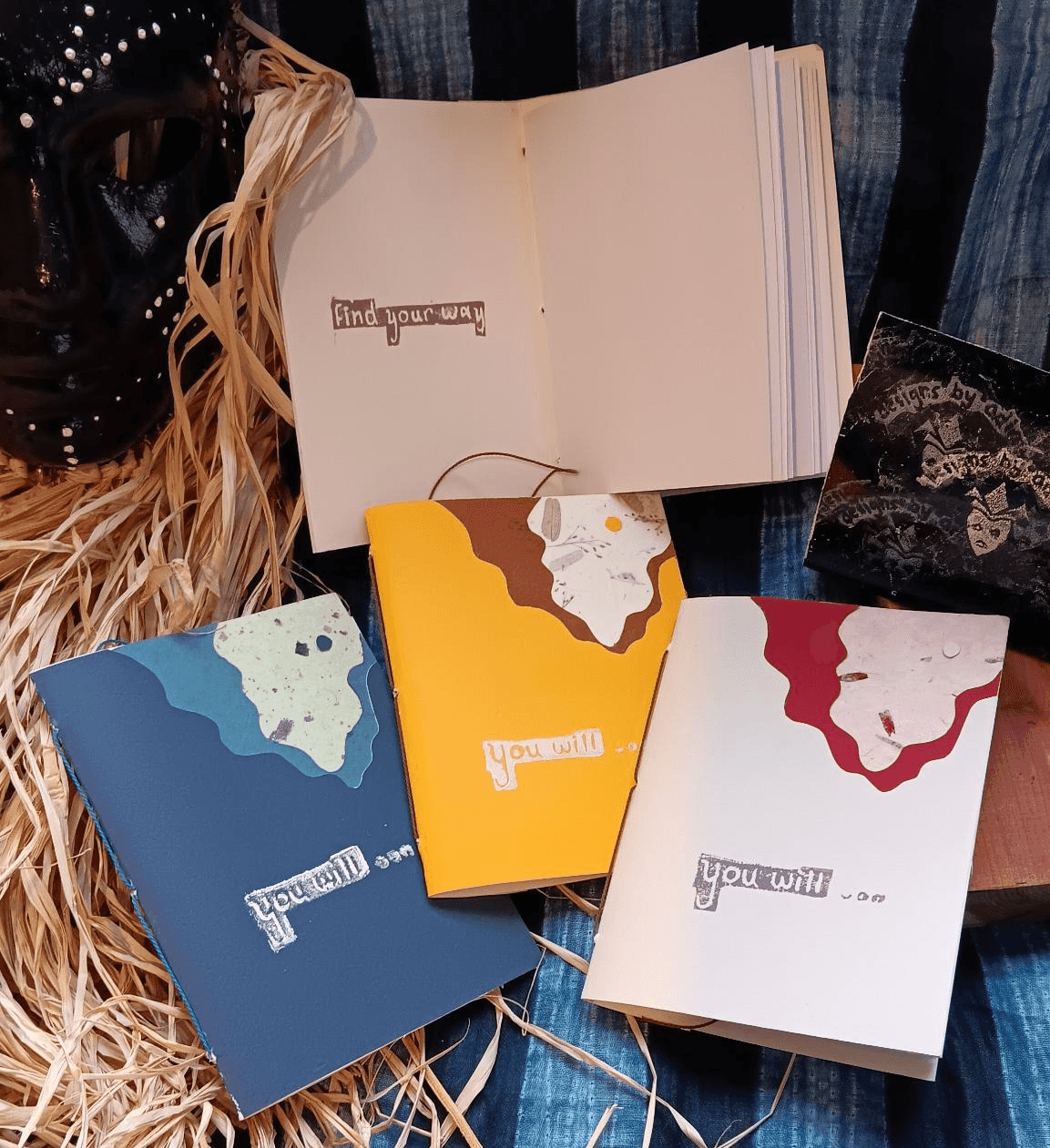 A couple notebooks set next to each other. The cover of the notebooks features different paper fibers, some smooth, some rough, cut, formed, and layered to produce gentle hills that open to reveal moon and sky.